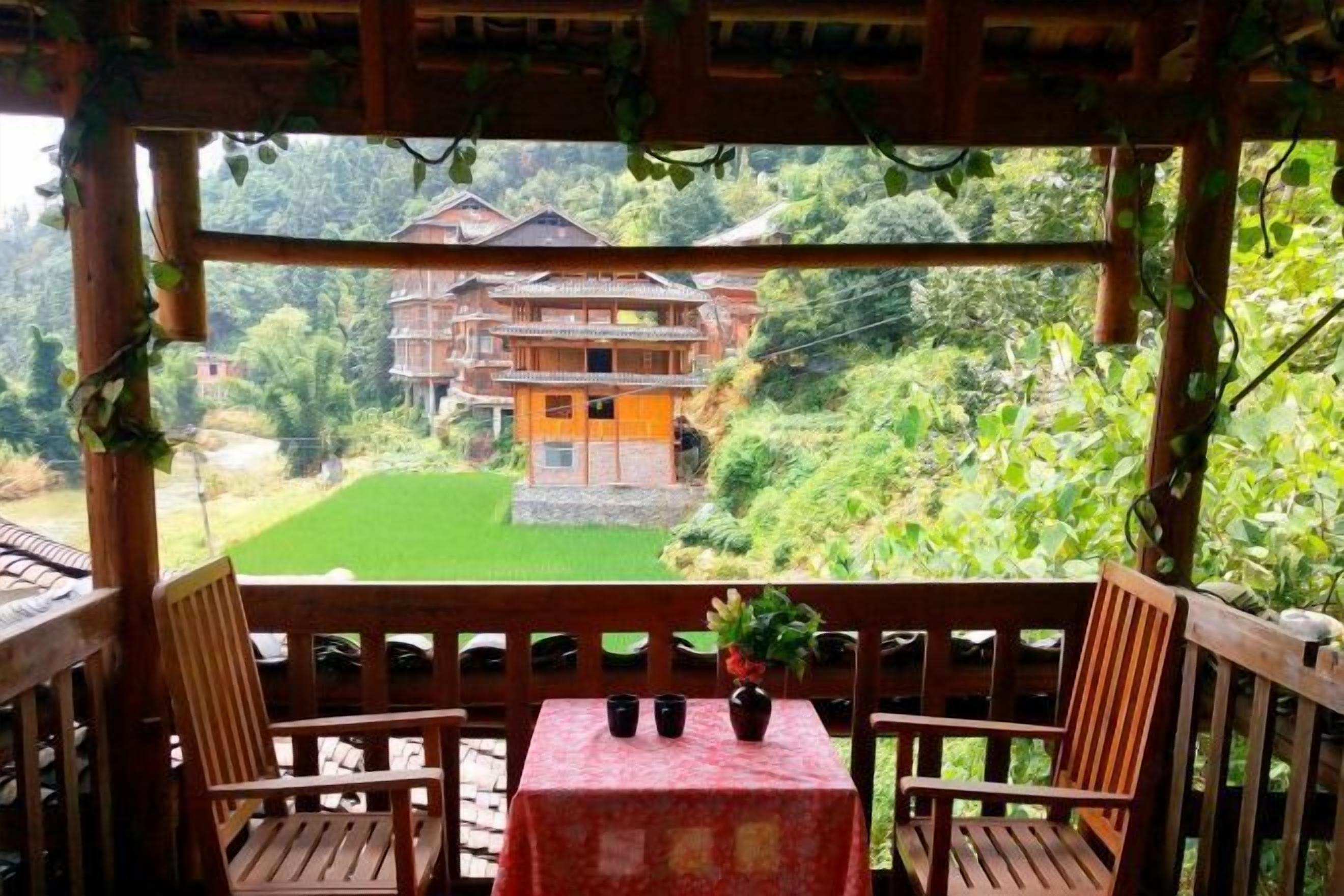 A wooden balcony with two chairs and a table, overlooking a lush green landscape and a traditional building.
