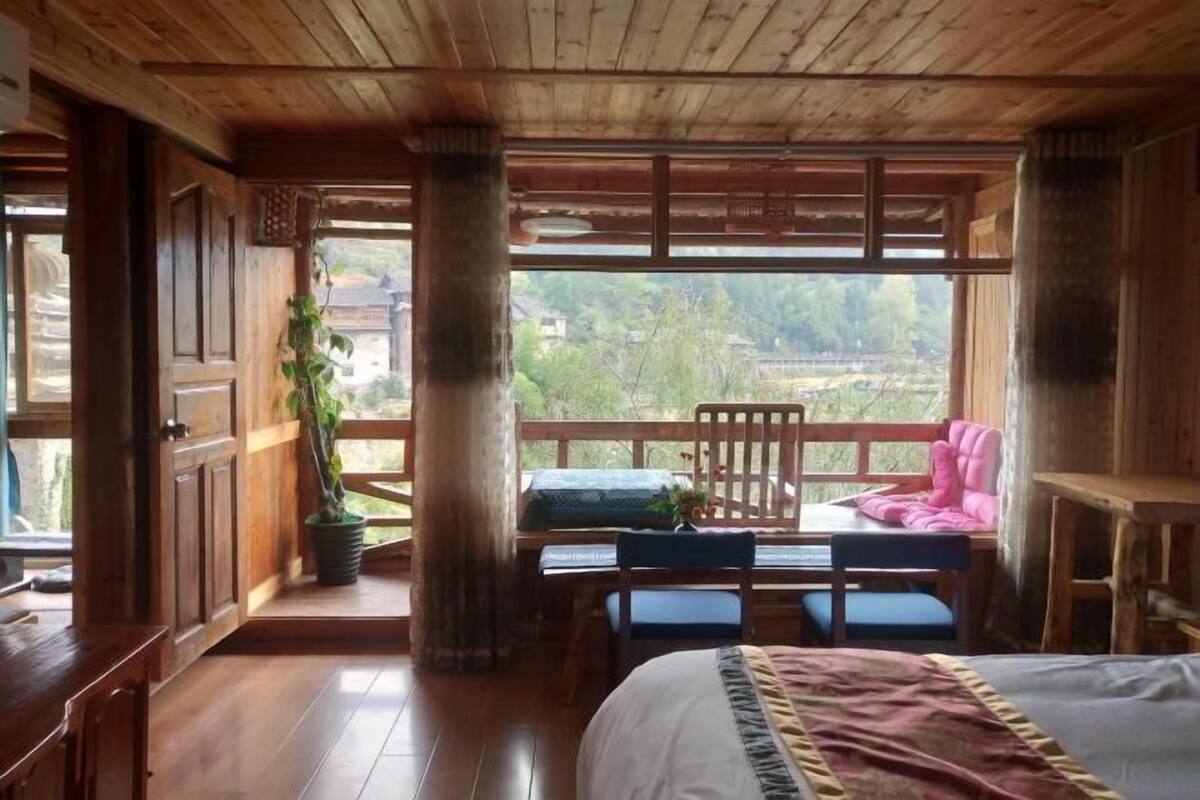 A wooden room with a bed, a desk, and a chair. There is a window with a view of greenery outside.