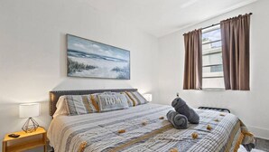Un dormitorio con una cama, una mesita de noche, una lámpara, una ventana con cortinas y un cuadro en la pared.