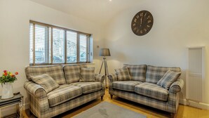 Un salon avec deux canapés à carreaux, un tapis gris, une horloge murale, un lampadaire et une fenêtre avec des stores.