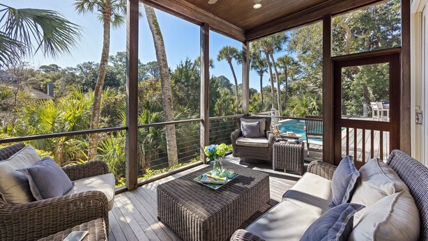 A covered outdoor seating area with wicker furniture, a ceiling fan, and a view of a pool and palm trees.