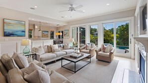 A spacious living room with a large sofa, a coffee table, and a dining area with chairs. There is a large window providing a view of the outdoors.