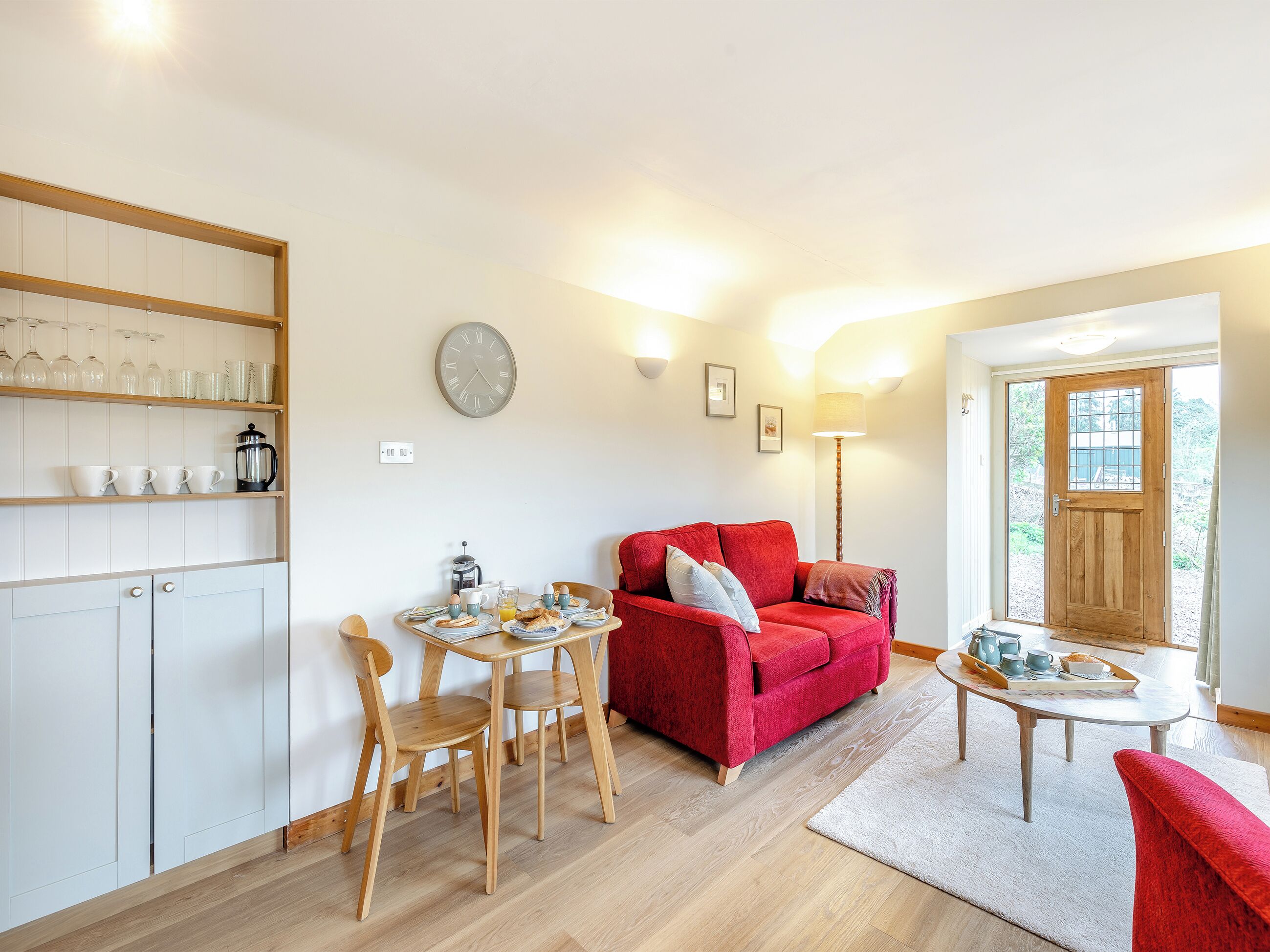 A cozy living room with a red sofa, wooden chairs, a coffee table, and a kitchen area with shelves and a clock.