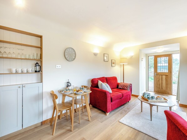 A cozy living room with a red sofa, wooden chairs, a coffee table, and a kitchen area with shelves and a clock.