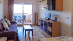 Ein Wohnzimmer mit einer Couch, einem Fernseher auf einem Schrank, einem Essbereich mit Stühlen und einem Tisch sowie einer Glastür, die auf einen Balkon führt.