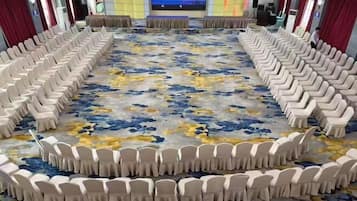 A conference room with rows of white chairs arranged in a semi-circle, a large screen at the front, and a carpet with a floral pattern.