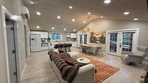 A spacious living area with a sofa, dining table, and chairs, a kitchen in the background, and a large painting on the wall.
