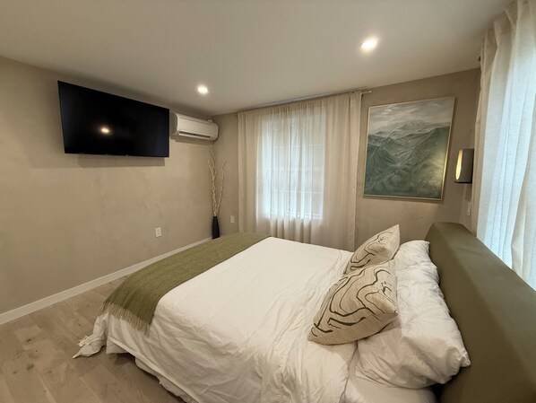 Un dormitorio con cama, televisor montado, equipo de aire acondicionado, un cuadro y una ventana con cortinas.