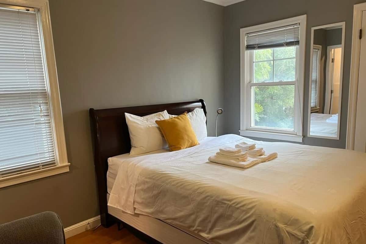 A neatly made bed with white linens and a folded towel, a wooden headboard, and a window with blinds.