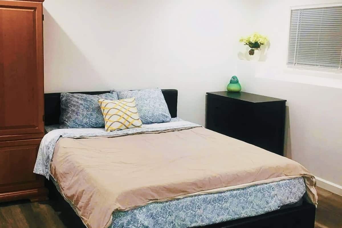 A bedroom with a bed, wooden wardrobe, bedside table, and a wall-mounted flower arrangement.
