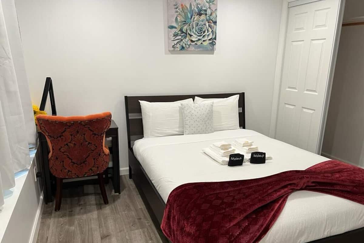 A neatly made bed with white linens and a red blanket, a chair with a patterned cushion, and a wall-mounted artwork.