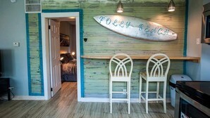 A room with a wooden wall, two white chairs, a surfboard sign, and a door leading to another room. - Boogie Down! Classic Folly Vibe, Lower level unit. (Folly Beach)