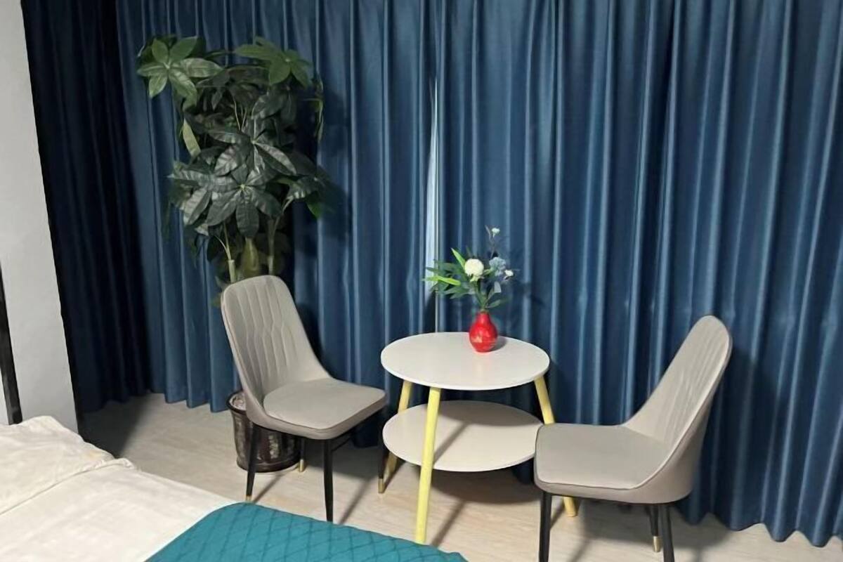 Una habitación con una cama, dos sillas, una mesita redonda con un florero y una planta grande en maceta.