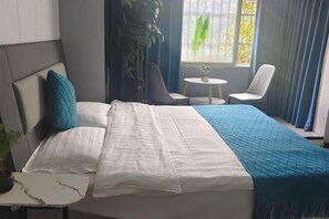 Ein ordentlich bezogenes Bett mit weißer und blauer Bettdecke, ein kleiner Nachttisch mit einer Pflanze und ein Fenster mit Vorhängen und Blick ins Grüne.