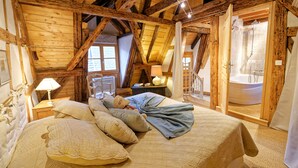 Een houten kamer met een bed, nachtkastje, lamp, teddybeer en uitzicht op een badkamer met een bad.