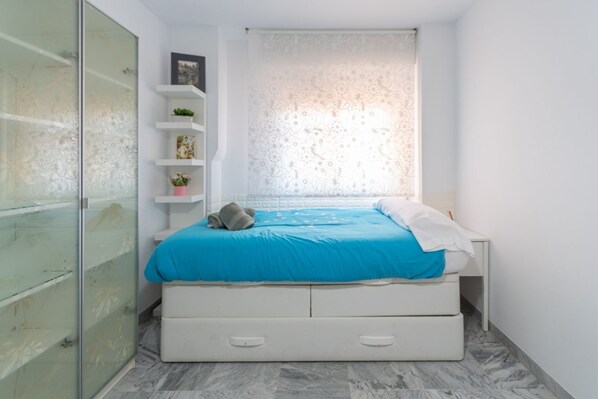 A bedroom with a bed, a nightstand, a dresser, and a window with a patterned curtain.
