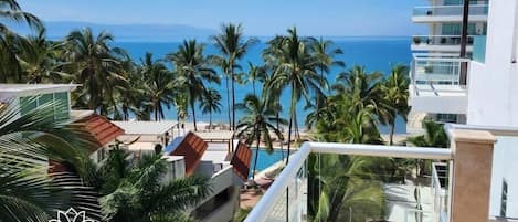 A high-rise hotel with a balcony overlooking a beach with palm trees and the ocean.