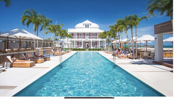 Un espace aménagé au bord de la piscine, avec des chaises longues et des parasols, une grande piscine et un bâtiment à la façade blanche et aux ornements rouges.
