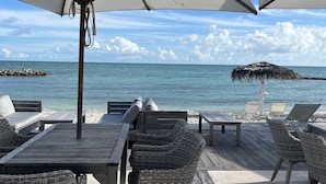 Une terrasse en bois, avec des chaises et des tables en osier, des parasols blancs et une vue sur l’océan.