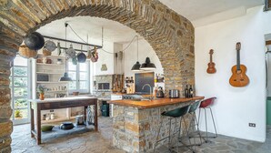 A rustic kitchen with stone walls, a central island, bar stools, hanging pots, and a guitar on the wall.