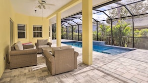 A covered patio area with wicker furniture, a pool, and a view of greenery.