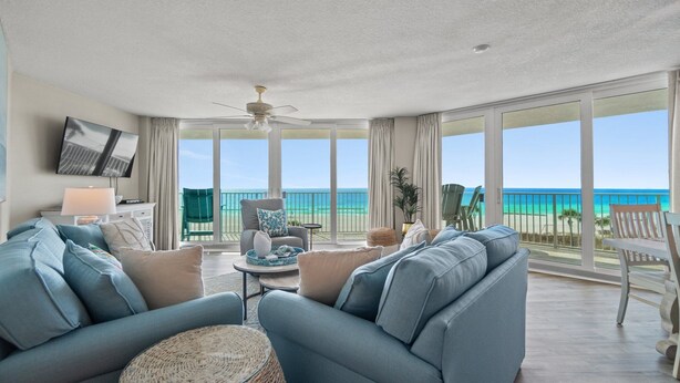 A spacious living room with a large window overlooking the beach, a ceiling fan, and a flat-screen TV mounted on the wall.