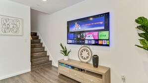 A modern living room with a flat-screen TV mounted on the wall, a wooden console table with decorative items, and a staircase leading to another level.