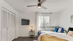 A bedroom with a bed, a ceiling fan, a bedside lamp, a window with curtains, and a TV mounted on the wall.
