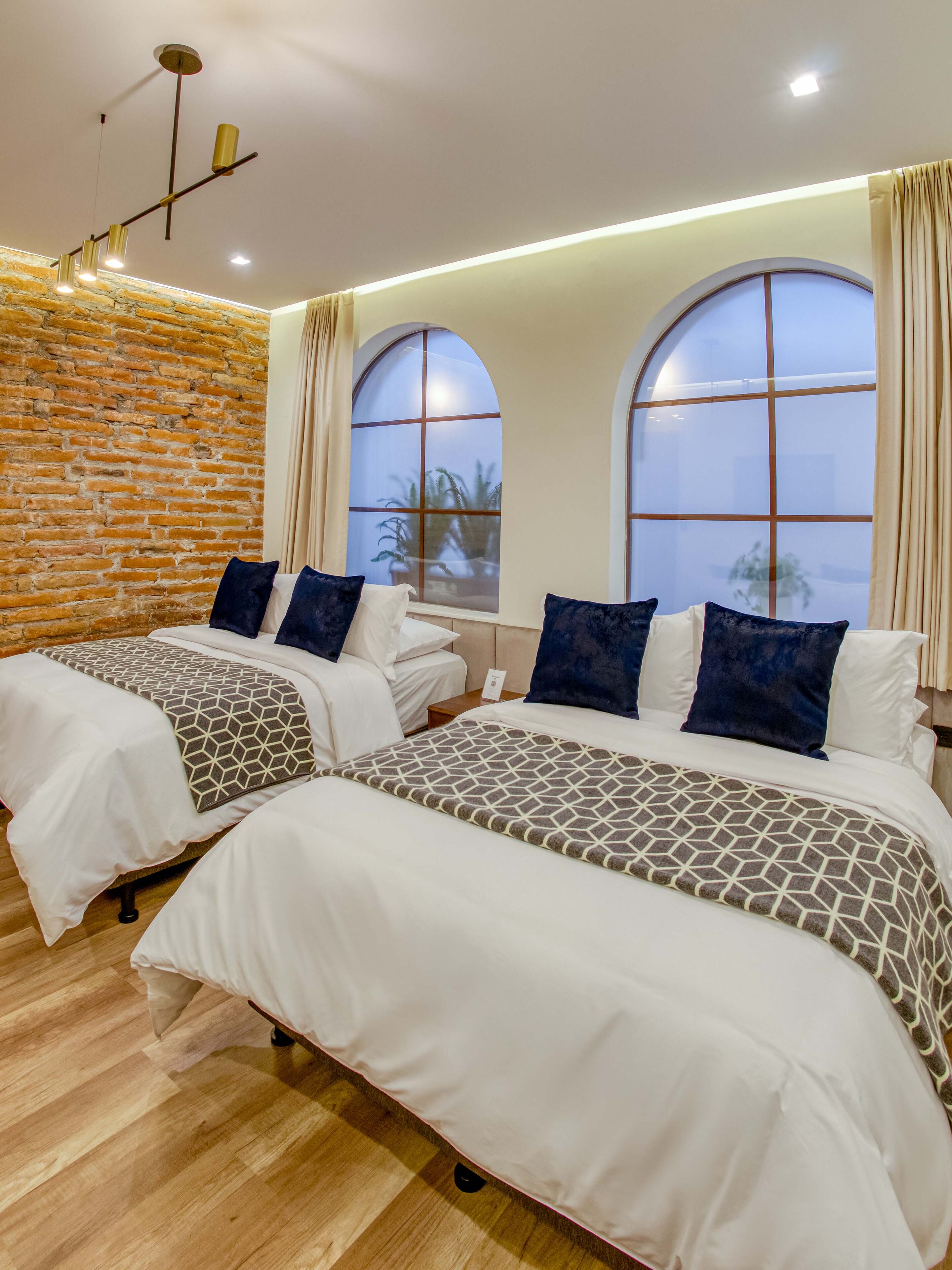 Camera d'albergo con due letti, parete in mattoni, ampie finestre ad arco e un lampadario moderno.