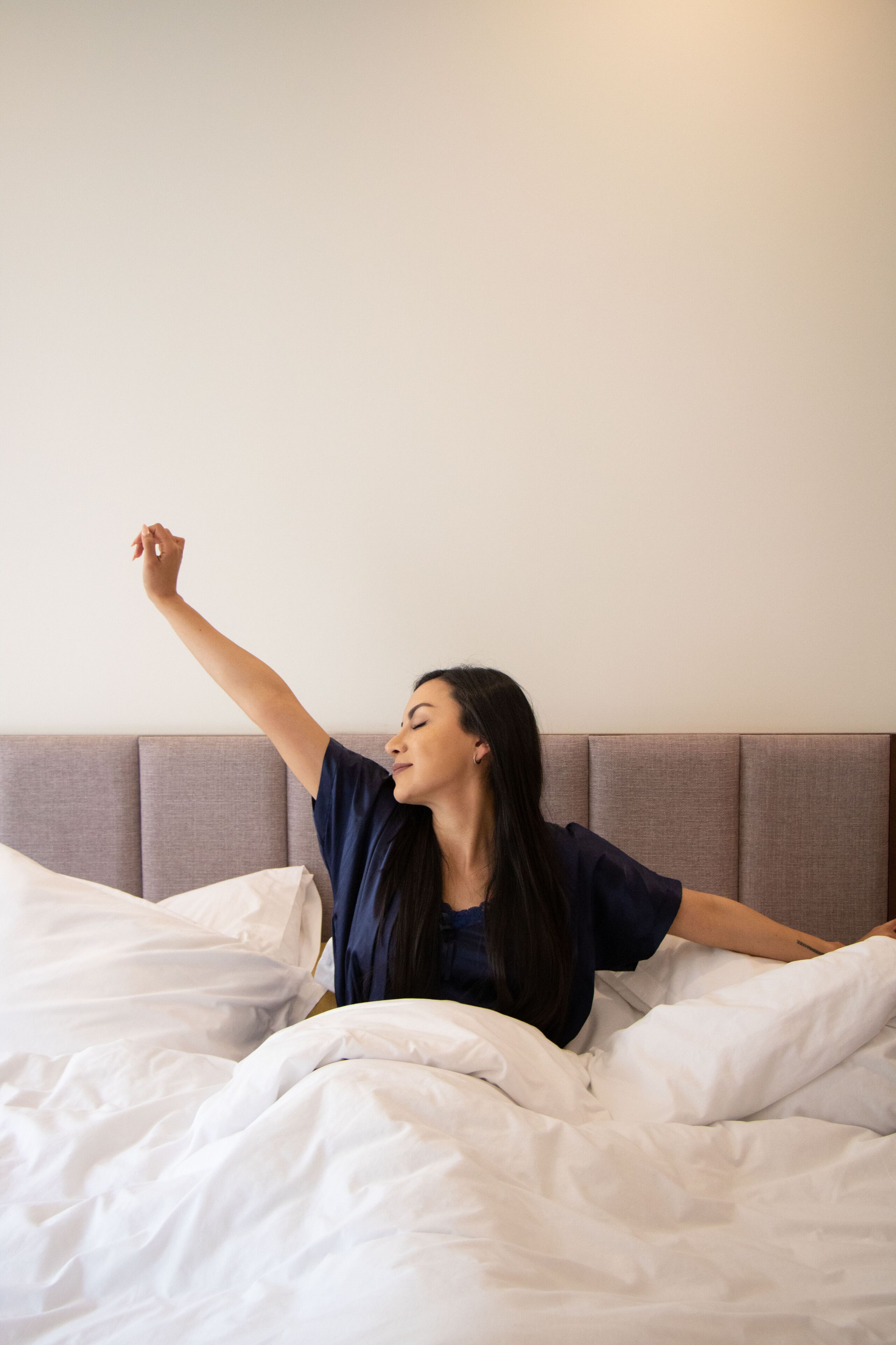 Una persona sentada en una cama con ropa de cama blanca y un cabecero gris.
