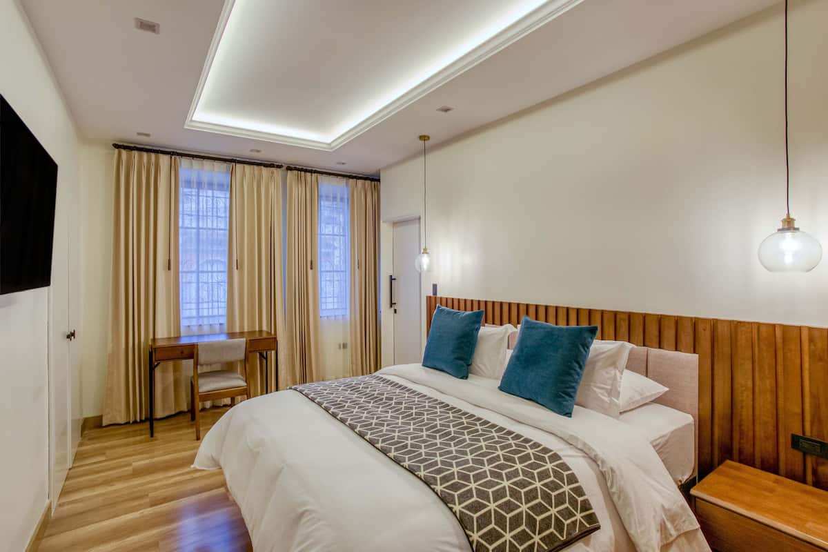 Une chambre d’hôtel moderne avec une tête de lit en bois, un grand lit avec une literie à motifs, un bureau avec une chaise et une grande fenêtre avec des rideaux.