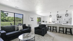 A modern living room with a sofa, armchair, and coffee table, a kitchen with white cabinets and bar stools, and large windows with a view of greenery. - Riverfront Ranch, Near Wine Tasting & Sedona (Cottonwood)