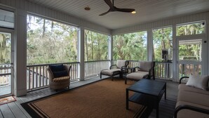 A covered porch with wicker furniture, a ceiling fan, and large windows overlooking a wooded area.