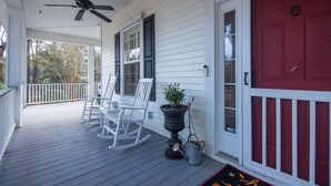 A porch with white rocking chairs, a potted plant, and a red door.