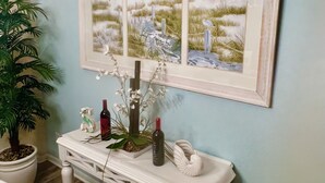 A room with a light blue wall, a white console table with a framed beach painting, a potted plant, and a white chair. - Villa Serenity (Davenport)