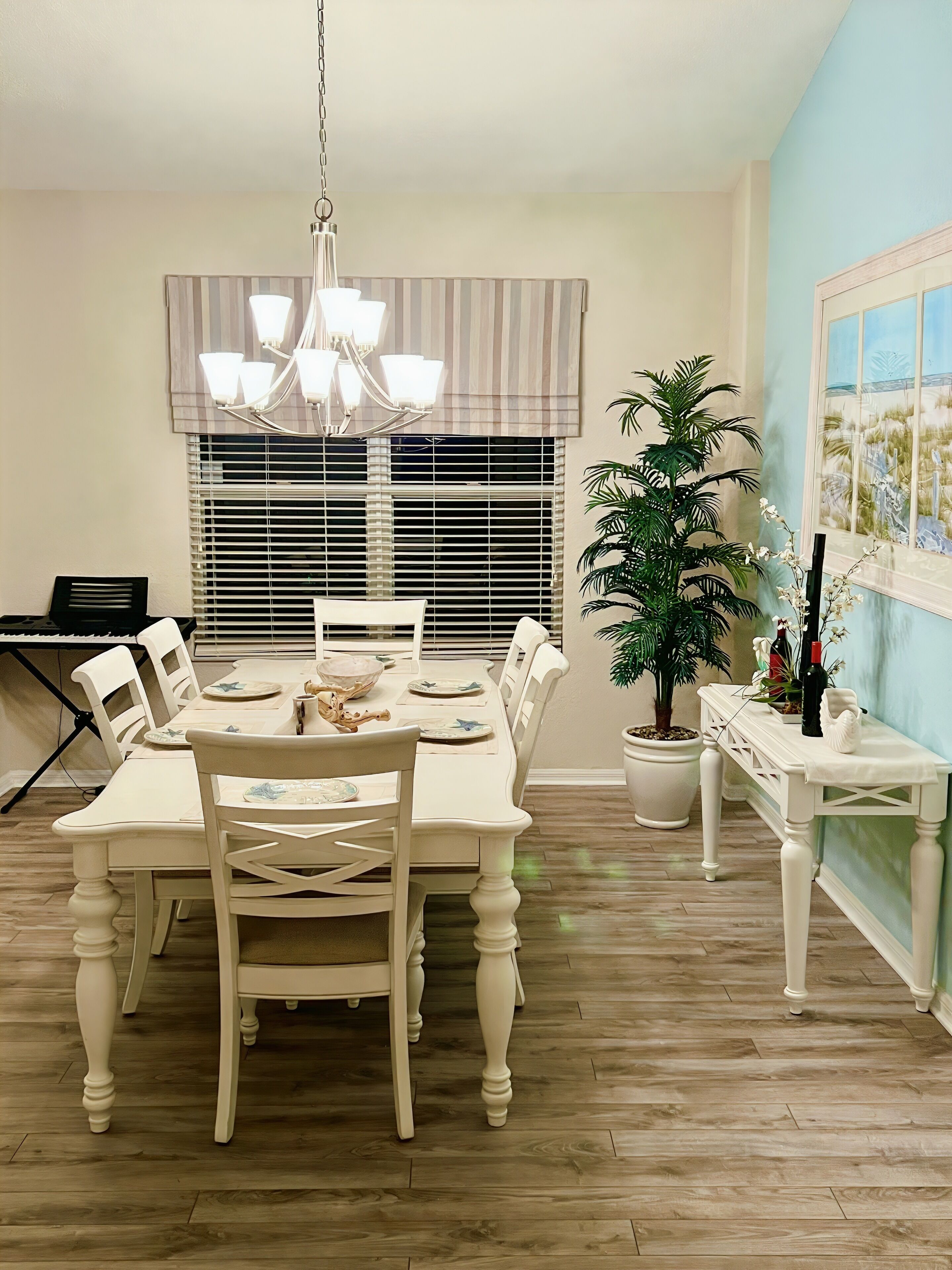 Un coin repas avec une table et des chaises blanches, un lustre, une plante en pot et une fenêtre avec des stores.