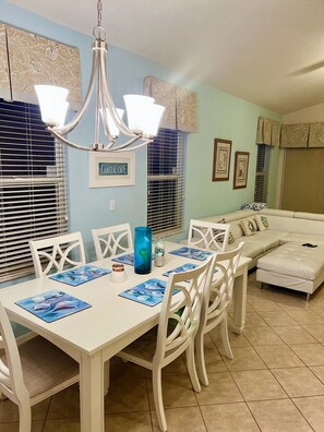 A dining area with a white table, chairs, and a chandelier. There is a sign on the wall and a sofa in the background.