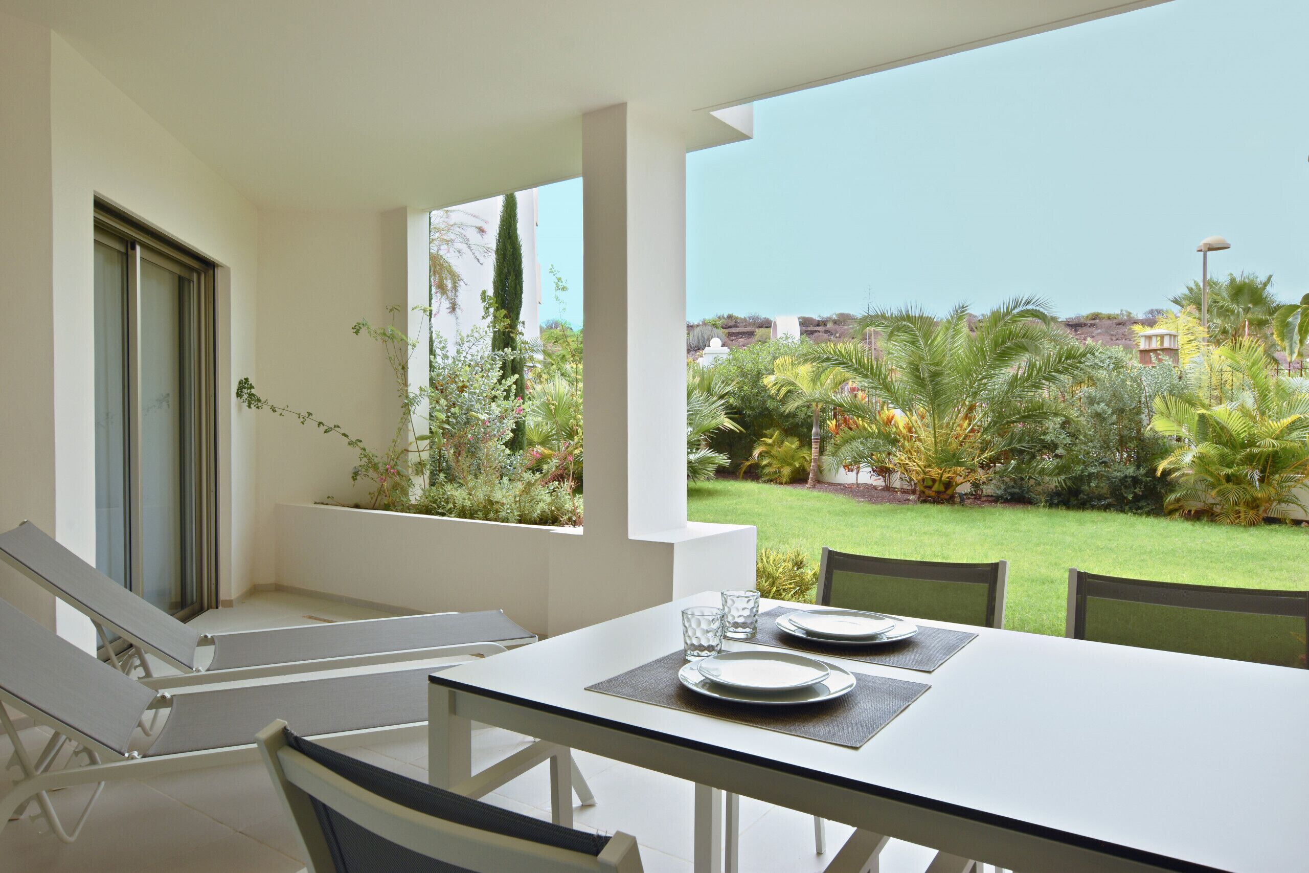 Un coin repas avec une table dressée pour deux personnes, une vue sur un jardin bien entretenu et un ciel dégagé.