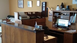 Un mostrador de recepción con una computadora, una impresora y un teléfono. Hay cuadros enmarcados en la pared y una estantería con varios objetos.