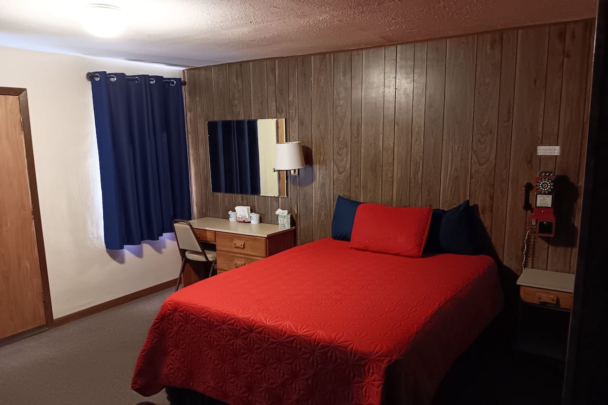 Ett hotellrum med en väggpanel av trä, ett skrivbord med en lampa, en säng med rött sänglinne, en telefon och ett fönster med gardiner.