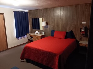 A hotel room with a wooden paneled wall, a desk with a lamp, a bed with red bedding, a telephone, and a window with curtains.