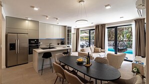 Una cocina moderna con una isla central, comedor con mesa y sillas, y un amplio ventanal corredero que da a una zona de piscina exterior.