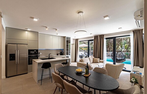 A modern kitchen with a central island, dining area with a table and chairs, and a large sliding glass door leading to an outdoor pool area.