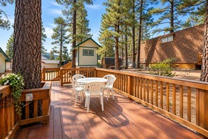 A wooden deck with a table and chairs, surrounded by trees and a building with a green roof.
