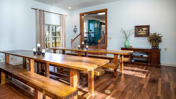 A spacious dining area with wooden tables and benches, a ceiling fan, and a view of a kitchen and staircase. - The Werken Haus | Pool | Pet-friendly | 2 Blks to Main (Fredericksburg)