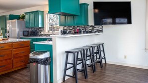 A modern kitchen with teal cabinets, a stainless steel refrigerator, a microwave, and a coffee maker. There are three bar stools and a trash can. - The Werken Haus | Pool | Pet-friendly | 2 Blks to Main (Fredericksburg)
