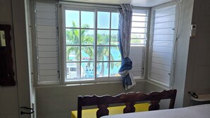 A bedroom with a bed, a window with a view of palm trees, a wooden chair, and a small table. - Sweets Beach View in the heart of  Ocho Rios (Ocho Rios)