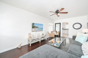 Ein modernes Wohnzimmer mit einem grauen Sofa, einem Glastisch und einer Holztruhe mit Dekoration. Ein Deckenventilator und eine Wanduhr sind zu sehen.