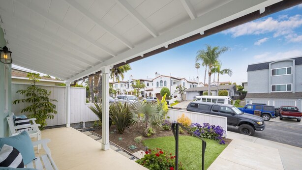 Un patio cubierto con mobiliario exterior, un pequeño jardín y vista a una calle residencial con coches estacionados y casas.