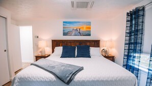 Un lit bien fait, recouvert d’une courtepointe blanche matelassée et agrémenté de deux oreillers, avec une tête de lit en bois, deux lampes de chevet et un tableau représentant un coucher de soleil sur une jetée.